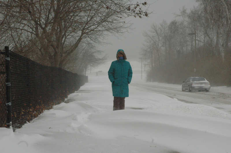 Una mujer vestida de verde parada en medio de la nieve. Al lado derecho hay un auto estacionado.